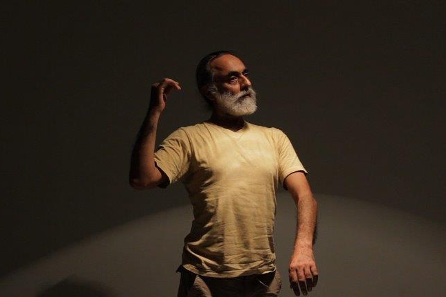 An image of Navtej Singh Johar, a man with a greying beard and moustache and black hair, wearing a white t-shirt. He is performing against a dark grey background with a spotlight on him. He is holding one arm up, bent at the elbow, and the other arm is swung out in front of him. 