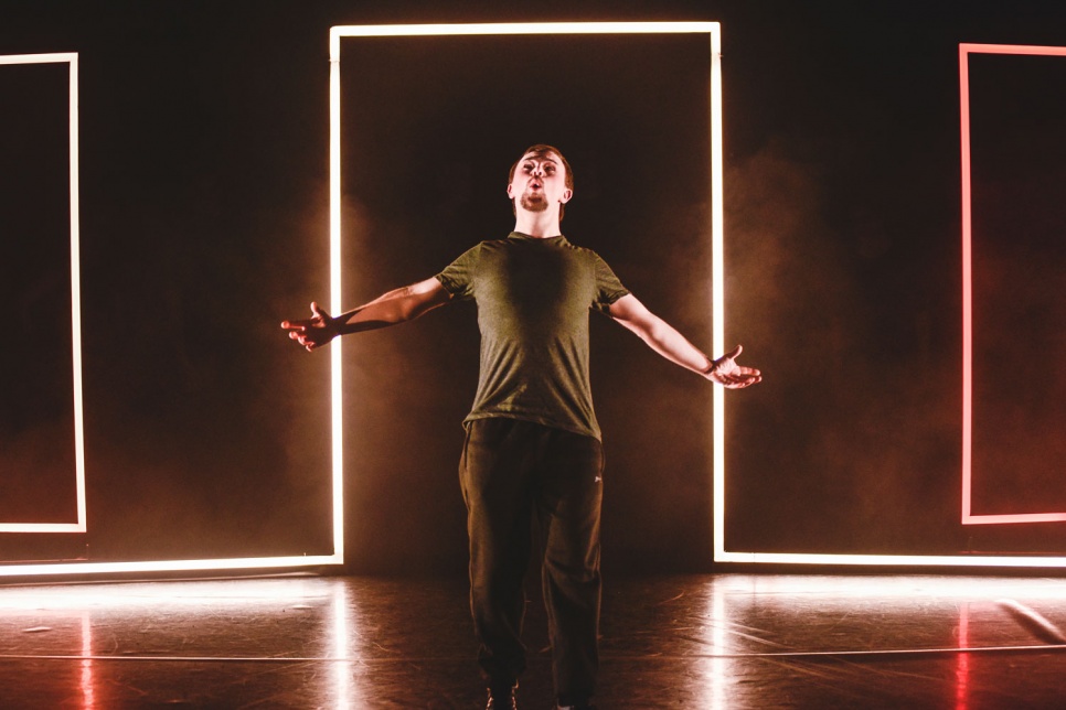 A young actor stands with arms outstretched on a stage with neon lights behind him