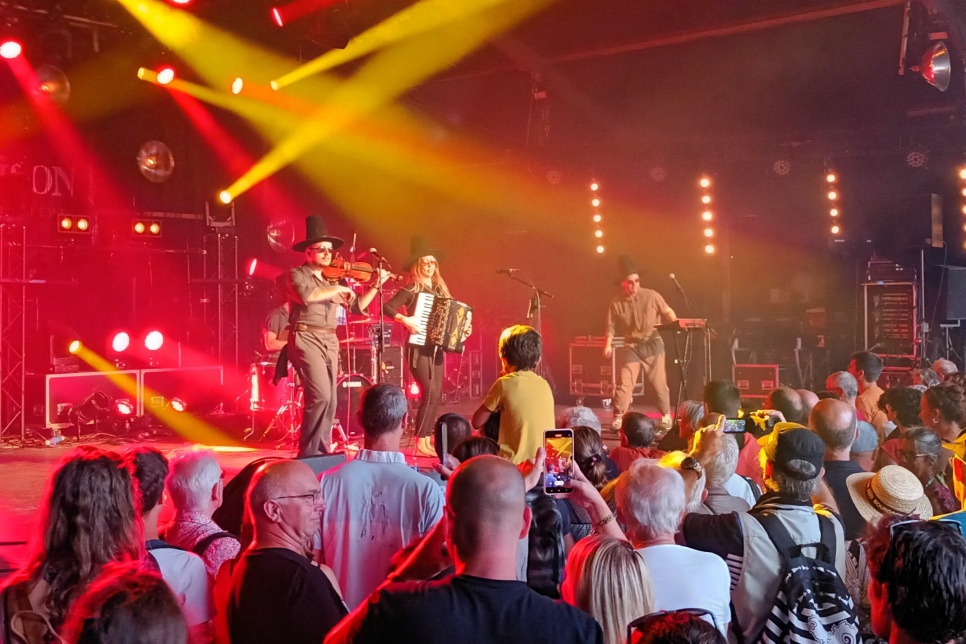 The band NoGood Boyo performing on stage with instruments to a large audience. There are yellow and red stage lights and the performers in the band are wearing traditional Welsh hats.