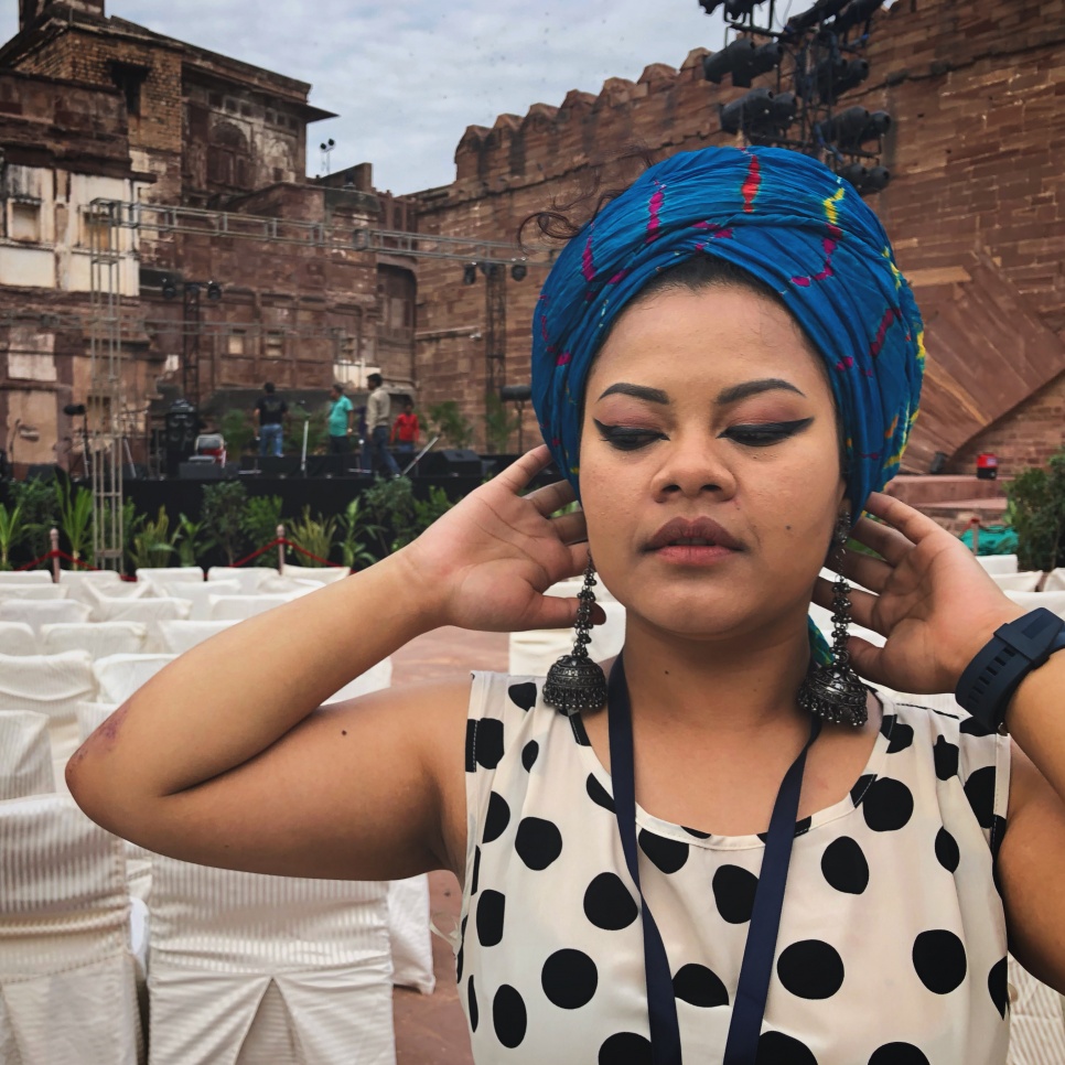 A woman in a black spotted dress and blue headscarf in front of the Mehrangarh Fort 
