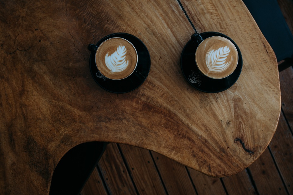 A birds-eye view of two black cups and saucers filled with coffee, topped with decorative coffee art, and sitting on a wooden table