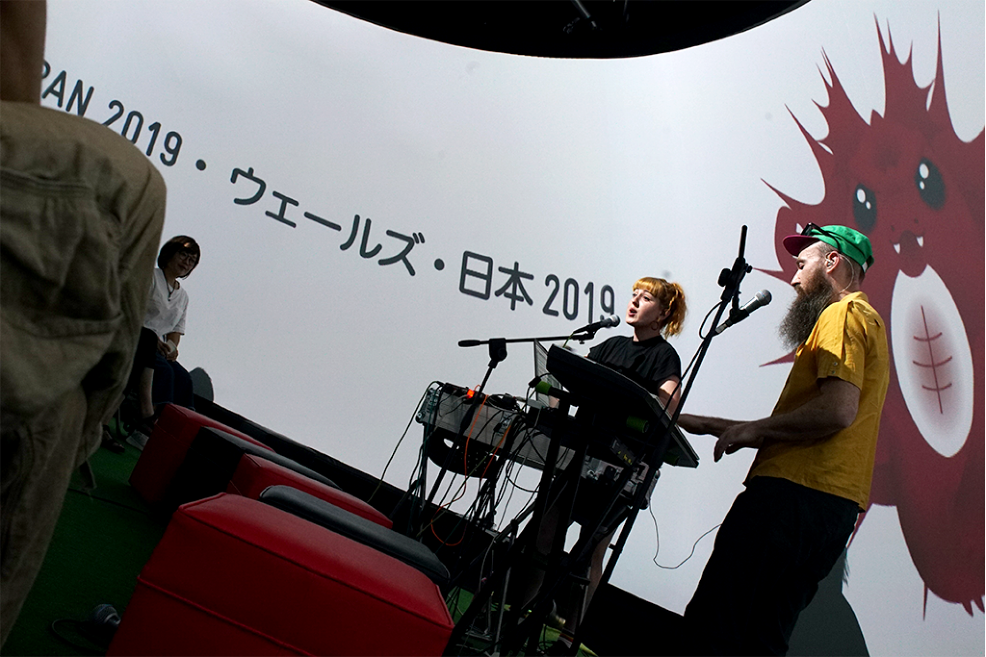 A male and female musical performance. Singing and playing keyboards in front of a large screen containing a cartoon character and Japanese writing