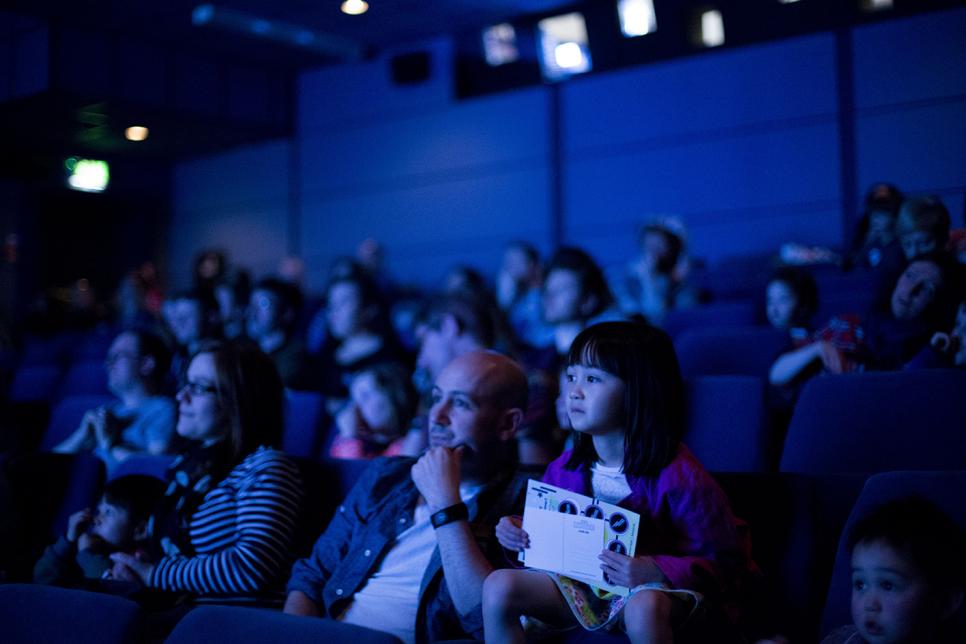 Audience members sit on seats looking at a screen out of shot.