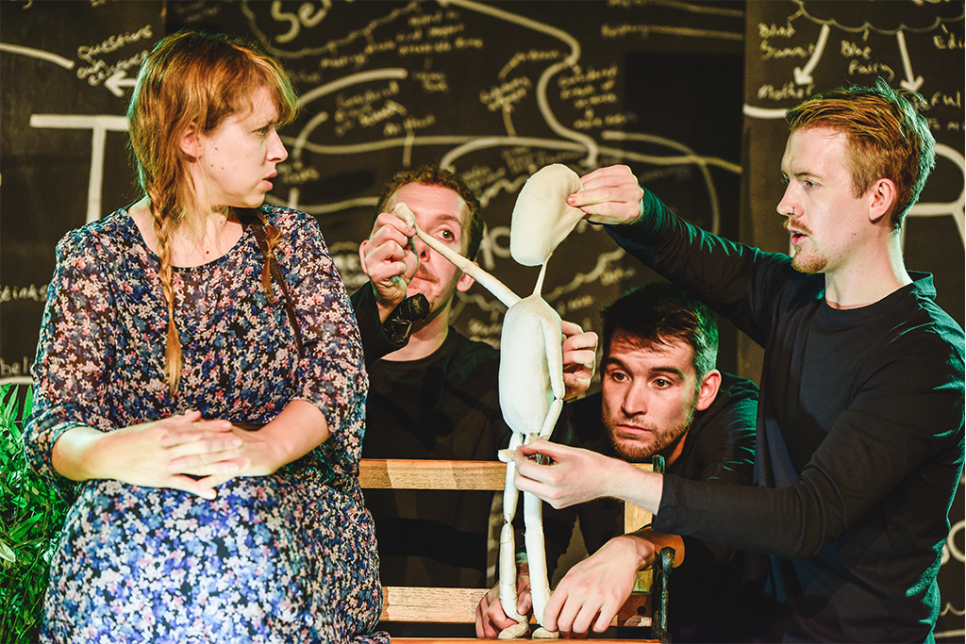 Four people acting onstage with a cloth puppet. Illustrations in the background