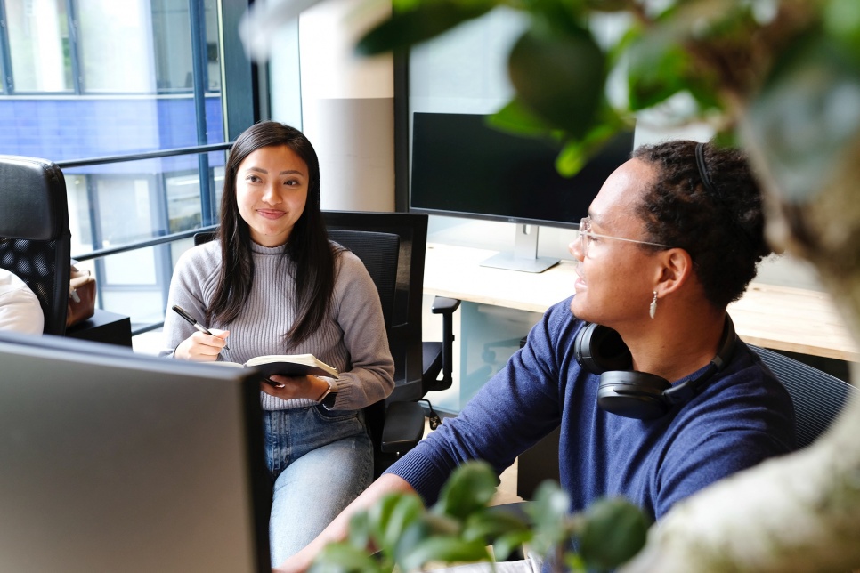 Two people around a computer. One wearing headphones and the other writing in a notebook