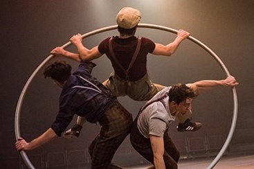 An image of 3 performers using a large metal ring wheel as a prop on stage 