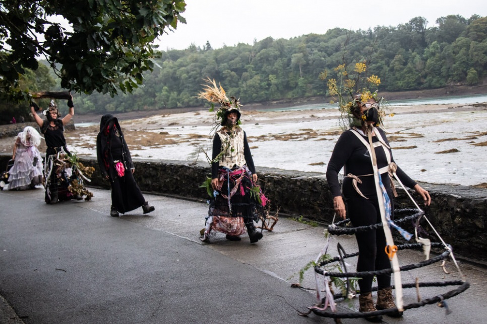 A group of performers with flower headpieces and consructed skirts walk alongside a river