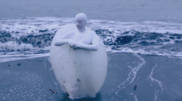 An artist dressed all in white is encased in a white balloon structure and is stood in the sea