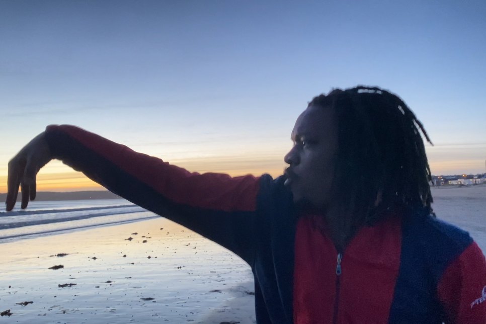 A dancer stands on a beach with their arm outstretched to the sea