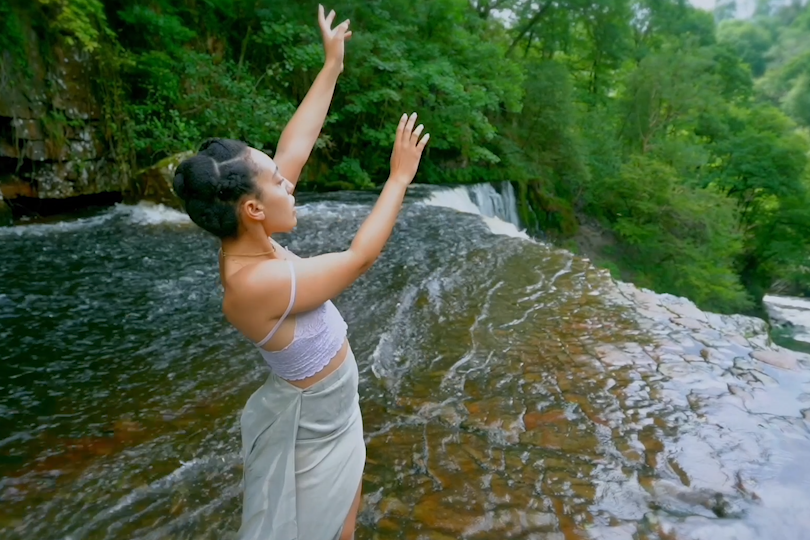 A dancer stands in a river with their arms to the air