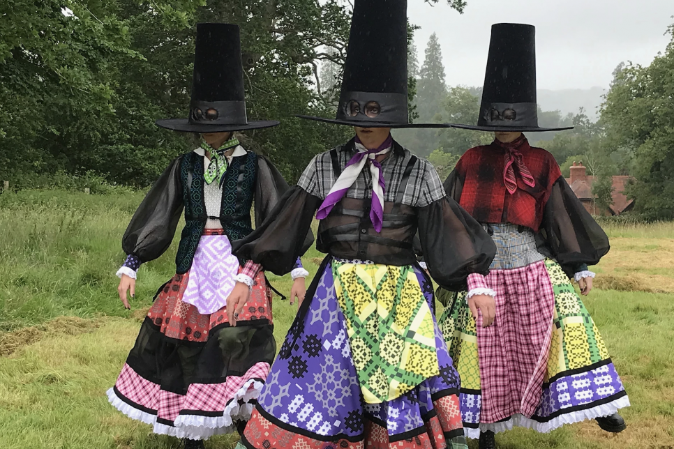 3 dancers in brightly coloured traditional Welsh costume, wearing extra large hats with eyeholes.