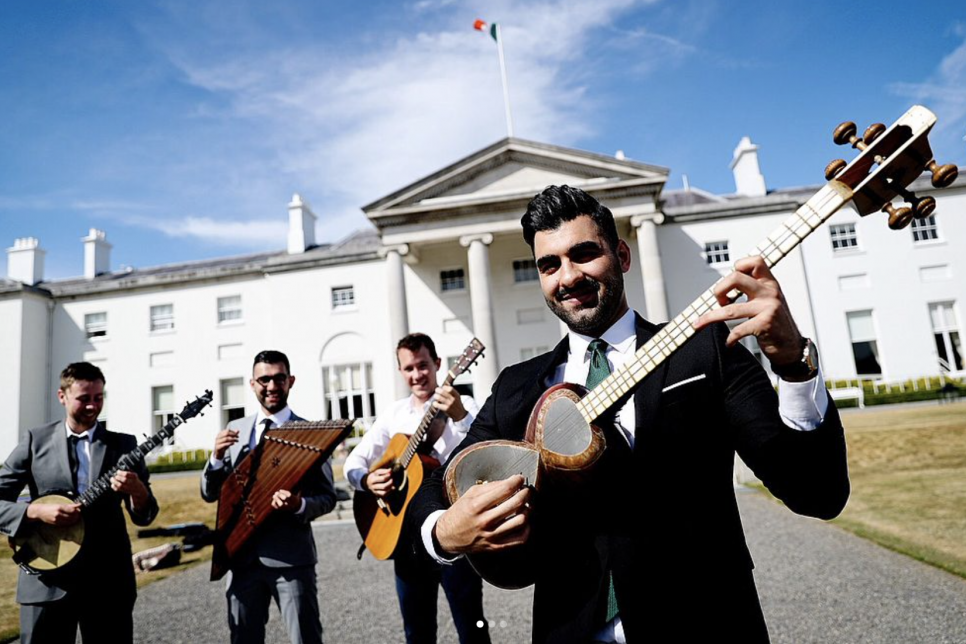 4 musicians in suits holding instruments stood outside of a large white stately building (Áras an Uachtaráin) 