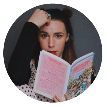 Mari Elen holding a hand to her face and a book in the other hand, in front of a grey wall.