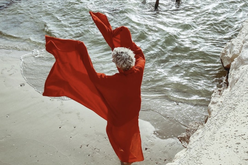 A person wearing a piece of red fabric which is flowing in the wind. They're stood on sand facing the water.