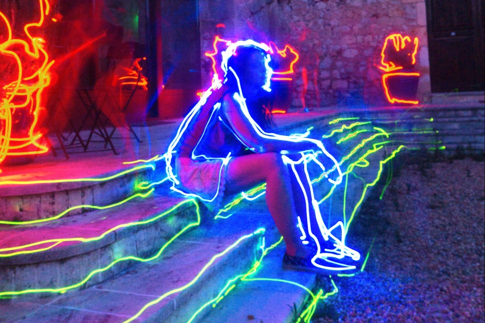 A person is sat on steps with the elbows resting on their knees. There are neon light leaks across the image tracing the outline of their body and the steps.