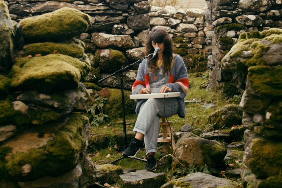 The artist Lisa Jên is sat in the middle of Wales Slate with mossy stones surrounding her. She has a small keyboard on her lap and is singing into a microphone.