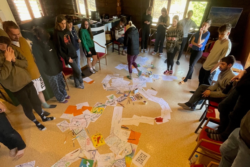 A group of people are stood in a circle looking at the floor where lots of paper is laid out. Some are stood on chairs and there is one person walking through the paper.