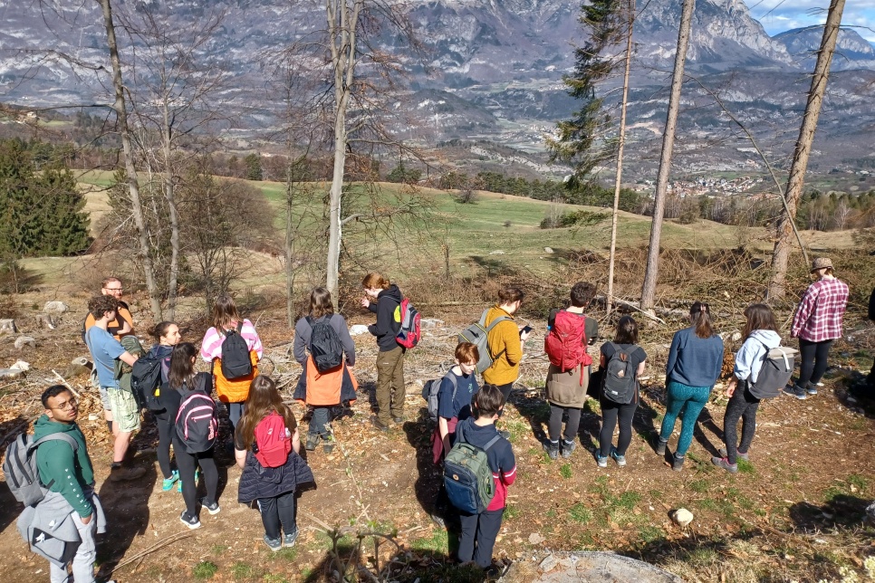 A group of people stood behind a line of trees looking out towards vast mountains. Most of the group are wearing backpacks and they are all gathered facing the direction of the mountain.