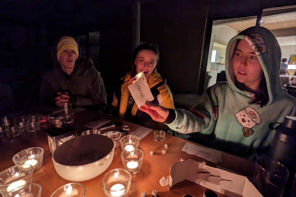 3 people sat at a table all in warm clothes. One person is holding up a piece of paper up which has been lit aflame, on the piece of paper it says 'insightful'. The other two look on at the burning piece of paper. There are a circle of lit candles on the table in front of them.