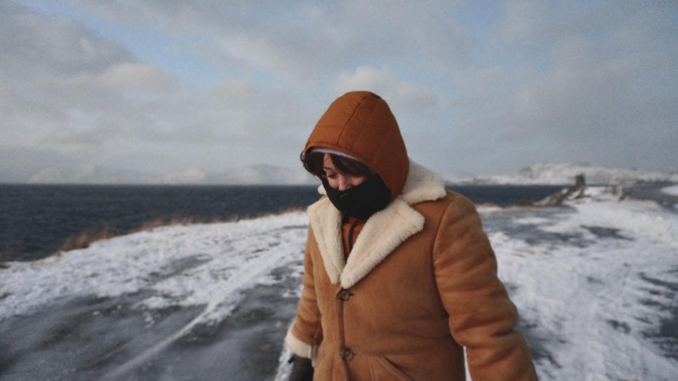 A person in a buttoned-up furry jacket and a scarf covering their mouth. They are stood centre frame as if walking towards the camera. Surround them is snow and ice covered ground, and the outer edge of a dark vast lake.