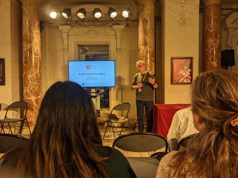 Two audience members are watching a speaker who is stood presenting next to a digital screen with a presentation slide that reads 'A new future for opera'. There are stage lights above the speaker and the surrounding room is grand with marble pillars surrounding the speaker.