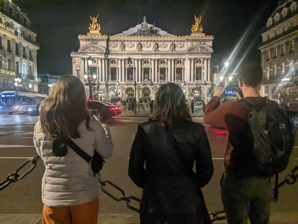 3 people are stood behind a chained barrier. They're looking at a grand building in the centre of Paris. The person on the left and the person on the right are holding their phones up to take pictures of the building.