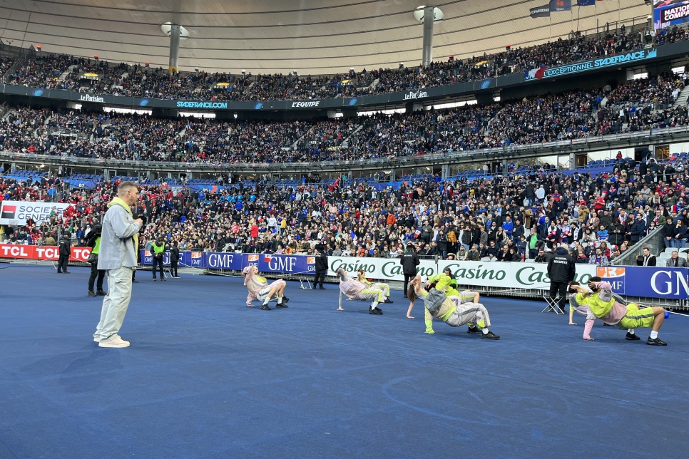 9 dancers are break-dancing on a blue carpeted area in front of a large crowd of people in the stands of the Stade de France