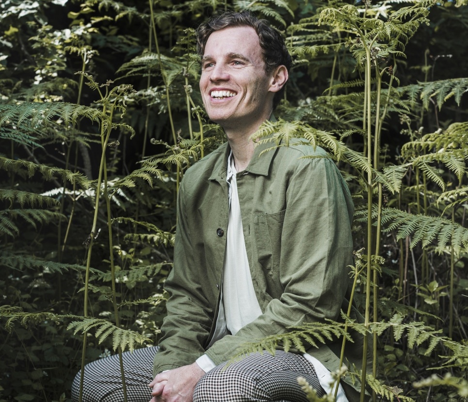 Man sat in green jacket, smiling, in front of foliage.