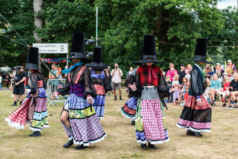 Performance of a group of people in bright, colourful dresses and Welsh hats, walking out in a circle.