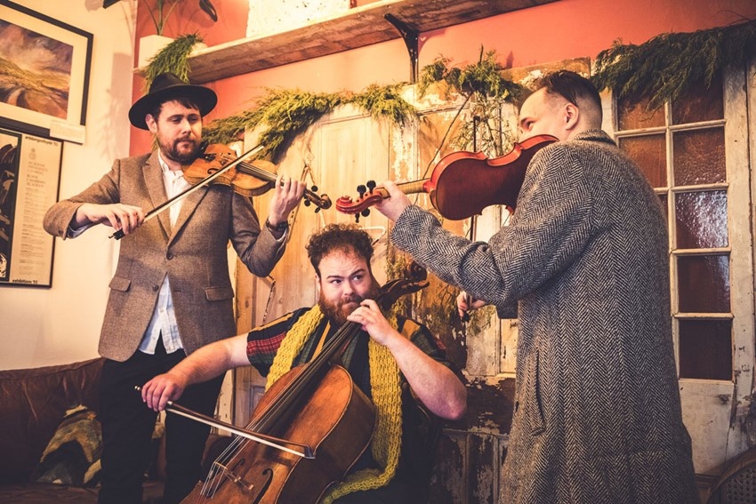 3 men in tweed jackets, playing string instruments in a cosy room setting.