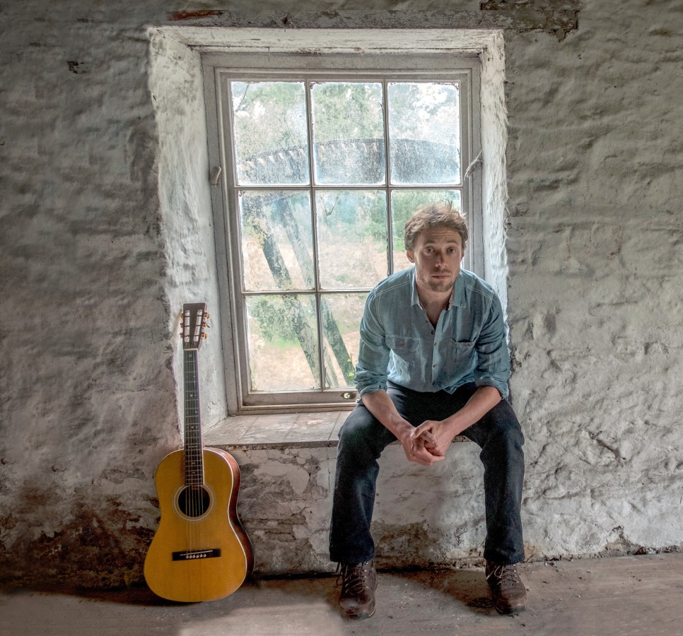 Man sat in a window in a blue shirt, next to a guitar.