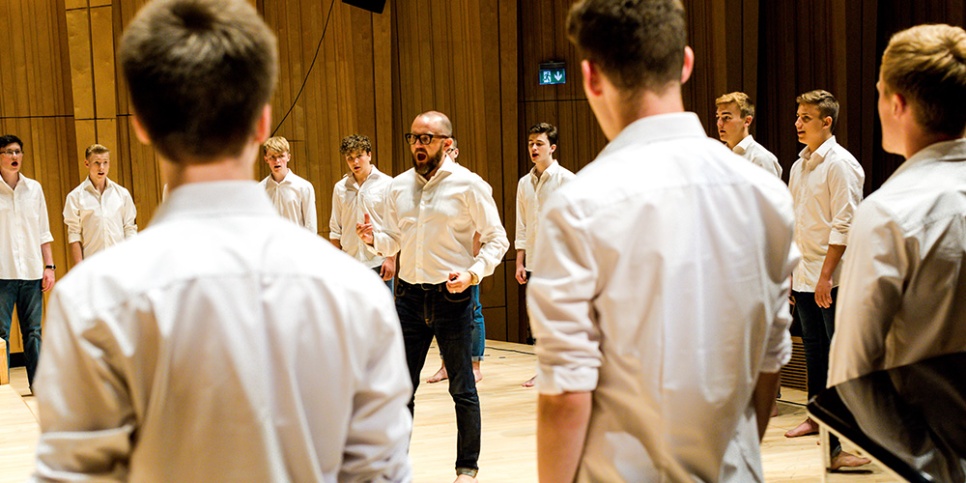 Group of men in white shirts, singing in a circle, in a wood panelled room.