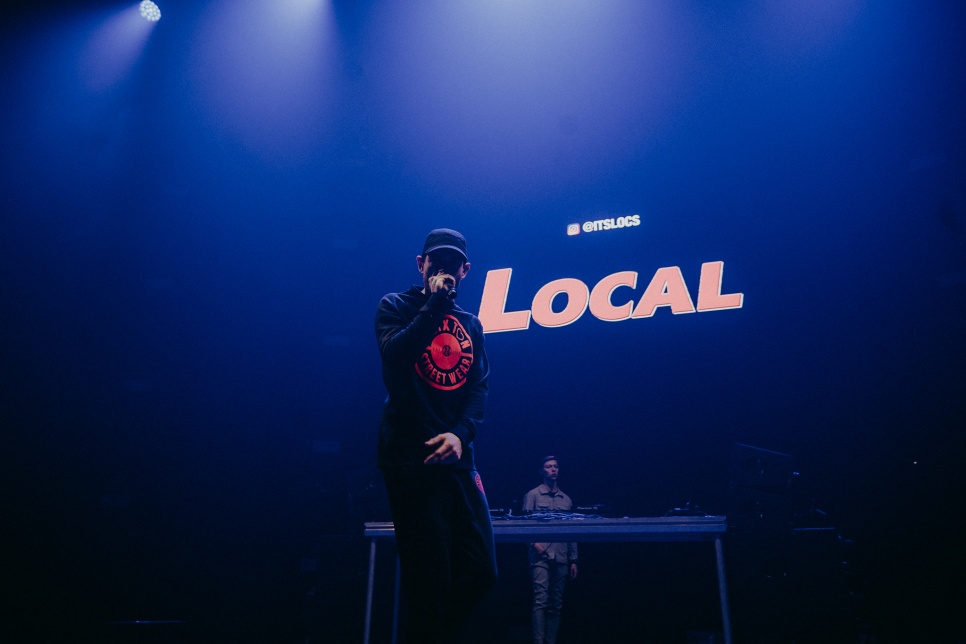 Man with microphone, dressed in black with black cap, on dark stage with the word 'Local' large in the background.