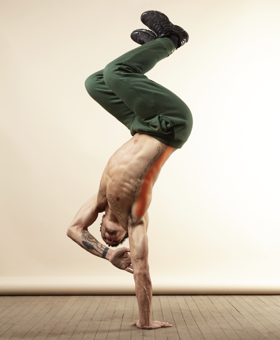 Man in green jogging bottoms, doing a handstand on one hand, in front of a light screen background.