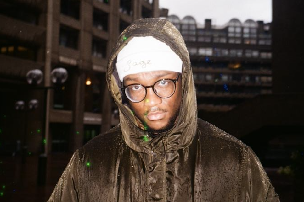 Man in front of a cityscape, wearing a brown hooded coat, white beanie and glasses.