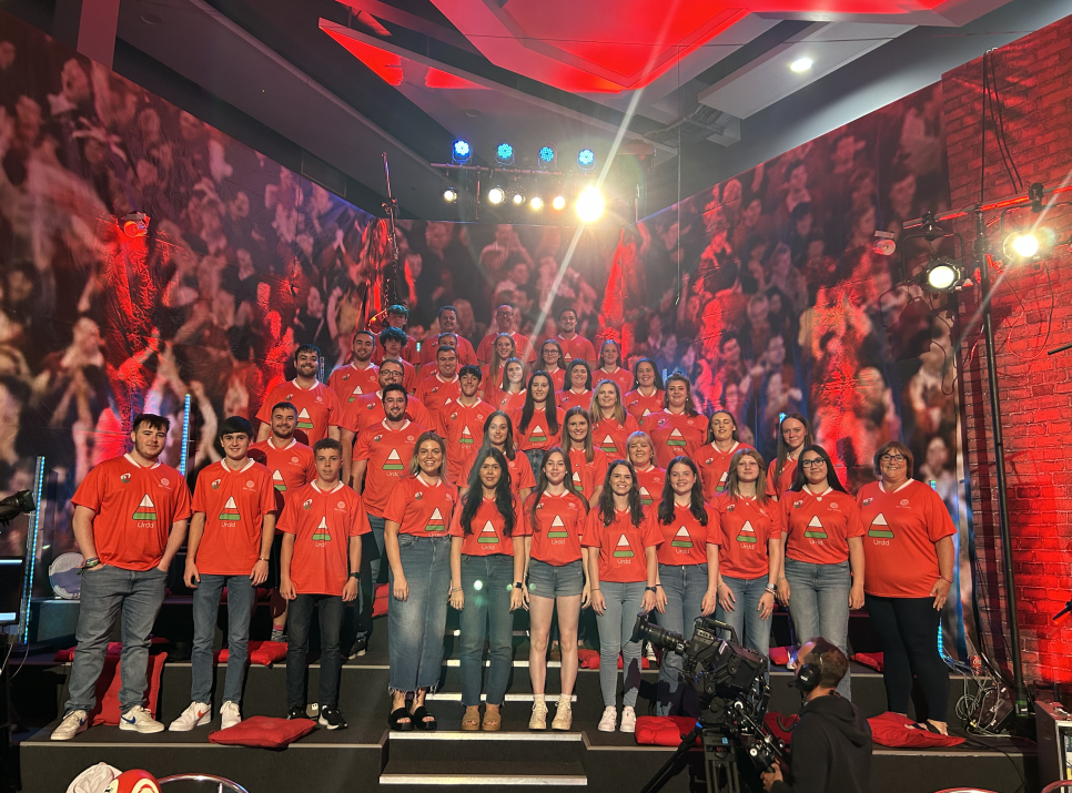 Large group of young people in red t-shirts stood in ascending rows, in a TV studio with a cameraman in the foreground.