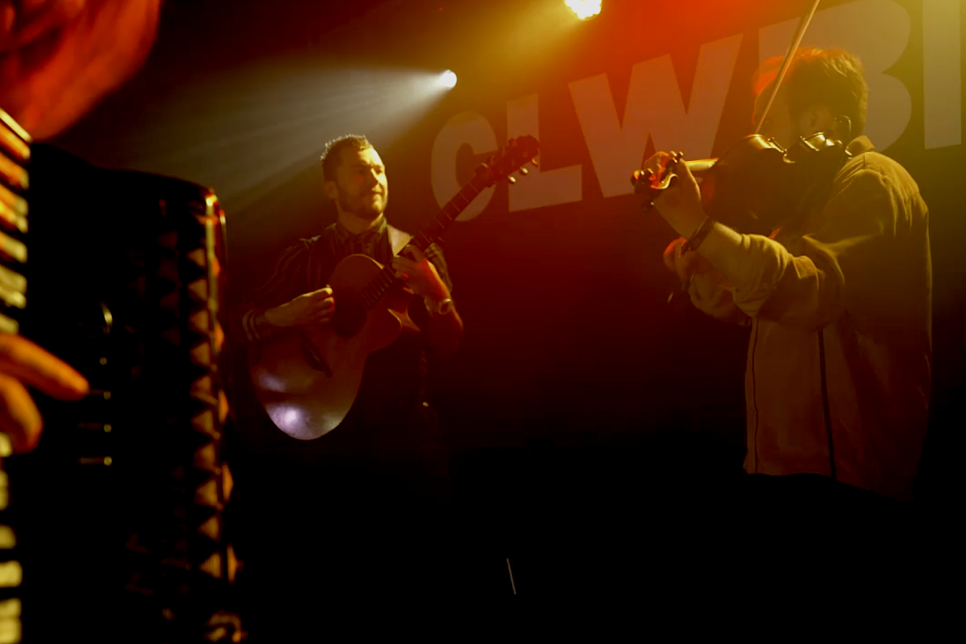3 musicians playing instruments on a dark stage with yellow and red stage lights.