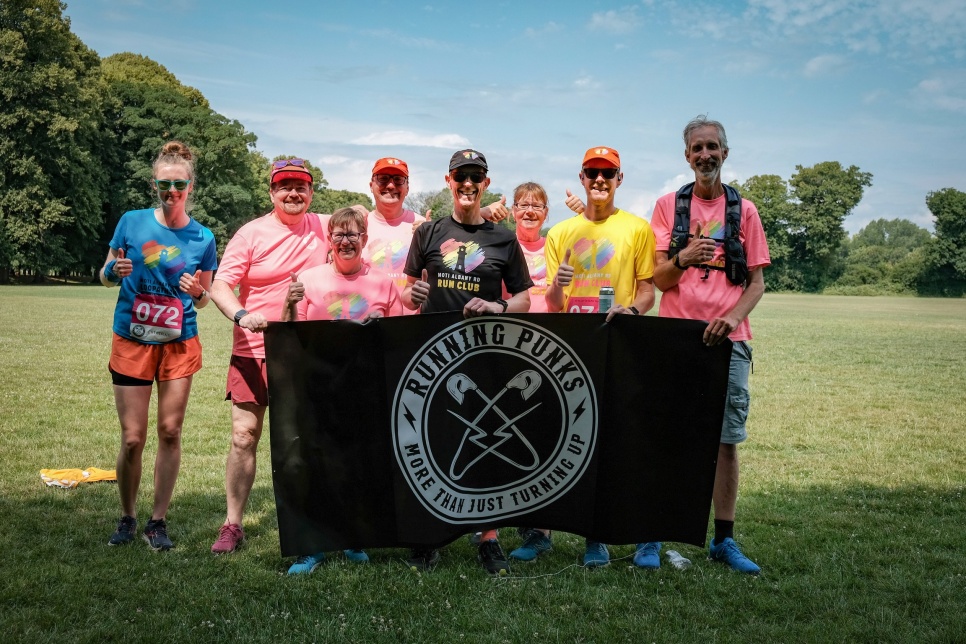 A group of people smiling in brightly coloured running clothing, on a sunny day in a field, holding a black 'Running Punks' flag.