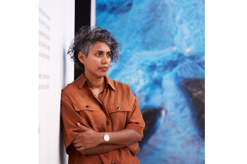A woman with short, wavy hair and brown shirt, with arms crossed, in front of bright blue artwork