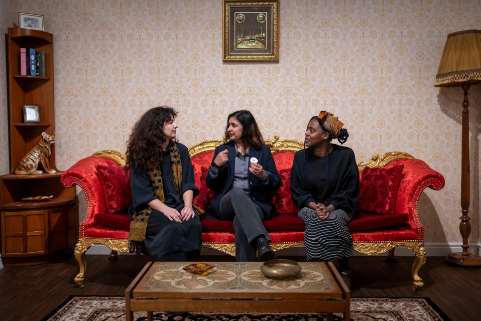 Artists Hannan Jones, Nasia Sarwar-Skuse, Jasmin Violet sit on a red vintage sofa once owned by Clive of India. A wallpapered backdrop featuring a framed portrait and a traditional lamp and various antique-style decorations complete the setting