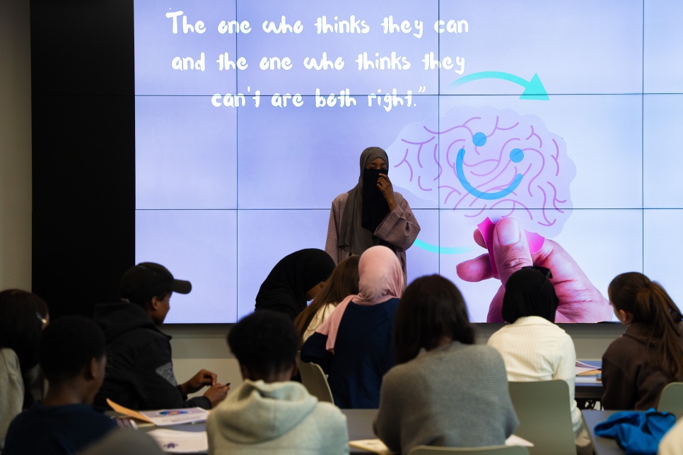 A room full of people with a woman presenting or performing at the font with a screen behind them. 