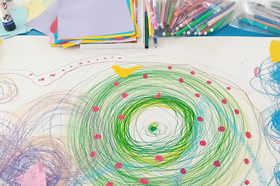 A colorful drawing featuring a large green spiral with small red hearts on a white paper, surrounded by crayons, markers, and assorted paper sheets on a table