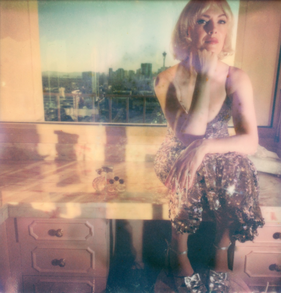 Women with short blonde bobbed hair,  sat in a sparkling dress in front of a sunny cityscape. 