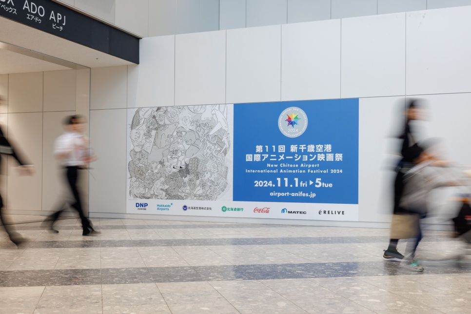 People walking in a white corridor by a large blue, white and black advertisement for an animation festival in Japanese text.