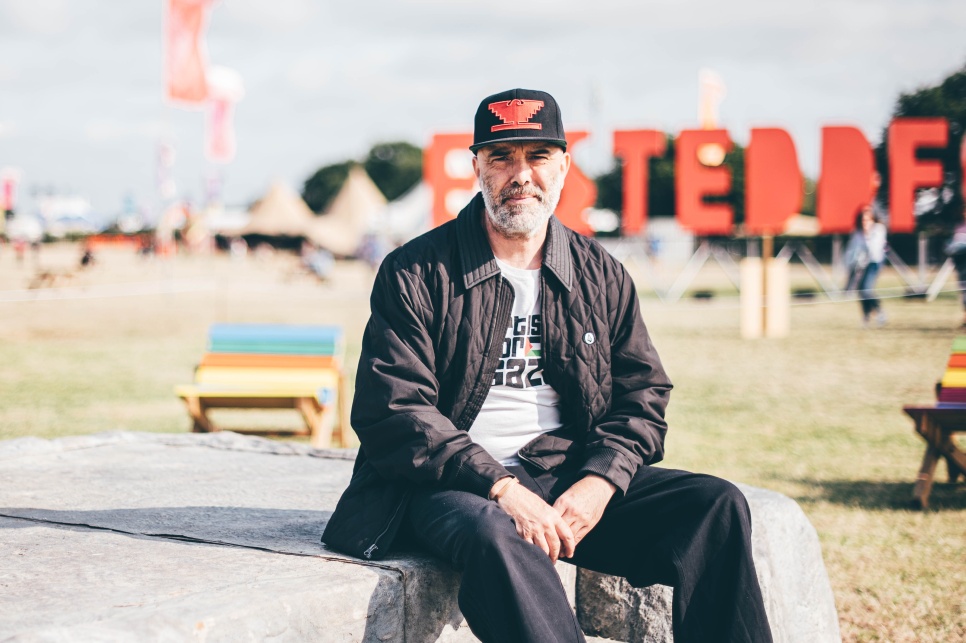 A man in a black jacket and black baseball cap sat on a large stone in front of a large red EISTEDDFOD sign
